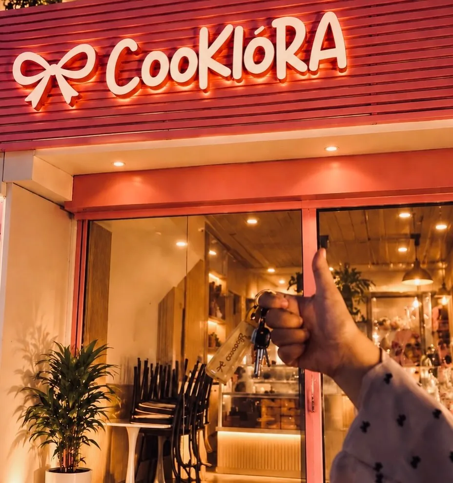 The Cookiora shopfront lit up, its bow and wordmark sign glowing, with a hand holding up the shop keys, thumb up.